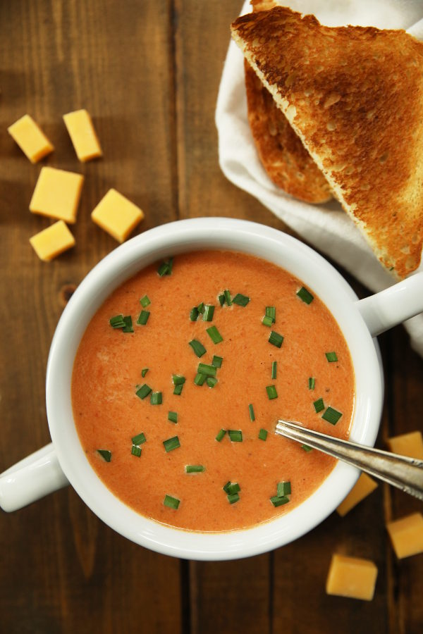 A white soup bowl with two handles is full of homemade tomato soup. The soup is a rich orange and garnished with some chopped chives. An old fashioned silver spoon is in the soup and chunks of cheddar cheese sit nearby as does a some toasted bread.