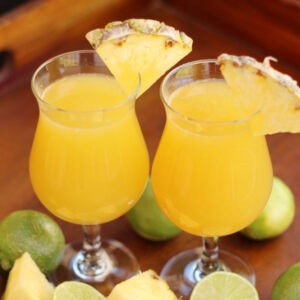Two glasses sit on a tray surrounded by chunks of pineapples and cut limes. The glasses each have a strong mai tai in them and a wedge of pineapple on the rim.