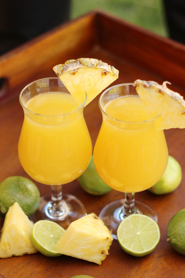 Two glasses sit on a tray surrounded by chunks of pineapples and cut limes. The glasses each have a strong mai tai in them and a wedge of pineapple on the rim.