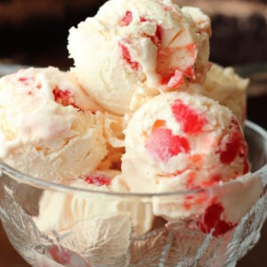 A clear glass dessert bowl holds big scoops of creamy amaretto ice cream. You can see large chunks of cherries in the ice cream. In the background there is a chocolate cake.