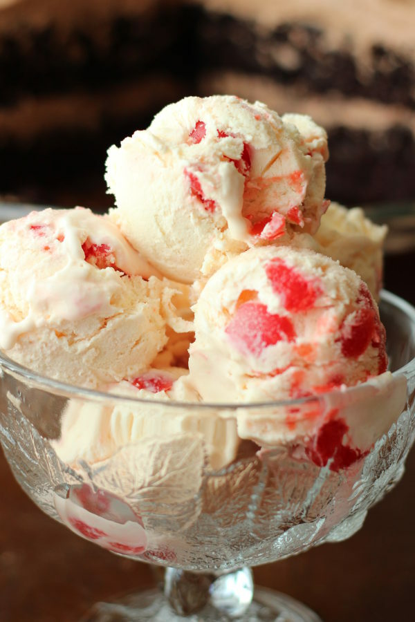 A clear glass dessert bowl holds big scoops of creamy amaretto ice cream. You can see large chunks of cherries in the ice cream. In the background there is a chocolate cake.