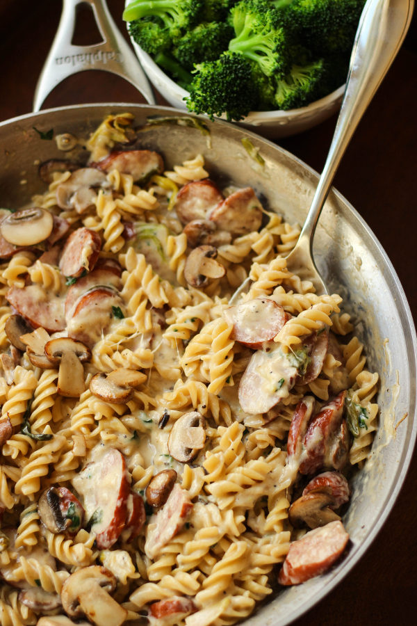 A large skillet is full of spiral noodles, Alfredo sauce, and slices of sausage. There is a bowl of steamed broccoli in the background.