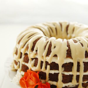 A luscious pumpkin bundt cake sits on a white plate. Cream cheese filling is between the layers and a brown sugar glaze is drizzled over the top. A few orange and white roses are at the edge of the cake.