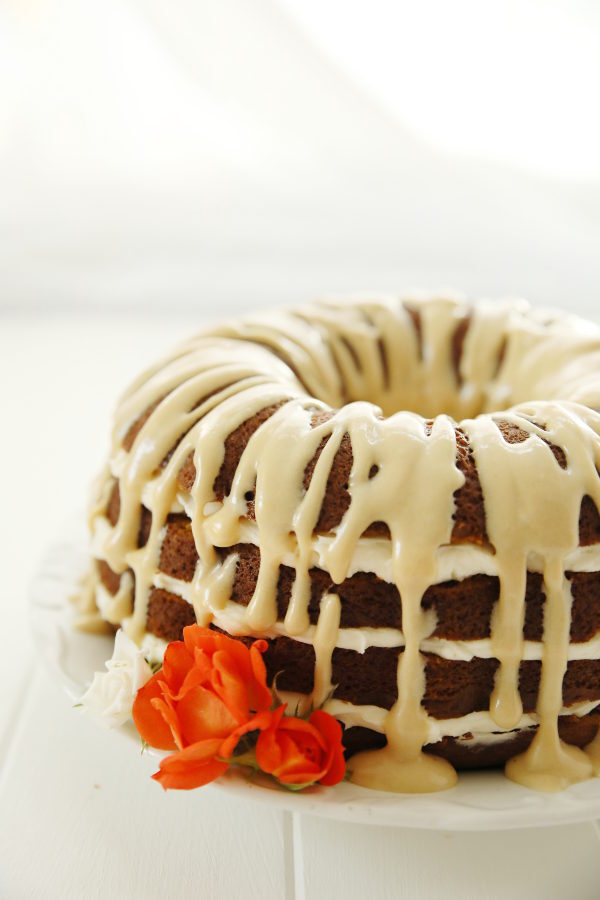 A luscious pumpkin bundt cake sits on a white plate. Cream cheese filling is between the layers and a brown sugar glaze is drizzled over the top. A few orange and white roses are at the edge of the cake. 