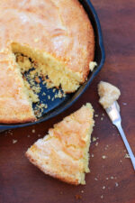 A cast iron pan is full of corn bread. The top is sparkling from a dusting of sugar and one wedge has been cut out and set on the table next to a knife full of honey butter.