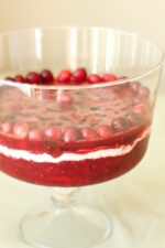 A large, clear trifle dish is half full of a cranberry jell-o salad. You can see beautiful ruby layers with cranberries, raspberries, and cherries. There is a layer of sour cream in the middle.