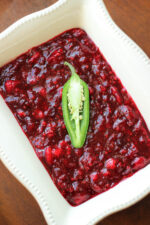 A large white rectangular container is full of cranberry, raspberry, jalapeno sauce. The sauce is crimson and chunky. A half a jalapeno pepper sits on top as garnish.