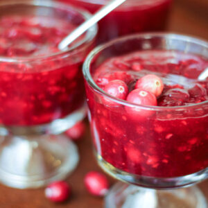 Three clear class bowls are full of crimson cranberry raspberry sauce. They have small spoons in them and a garnish of fresh cranberries.