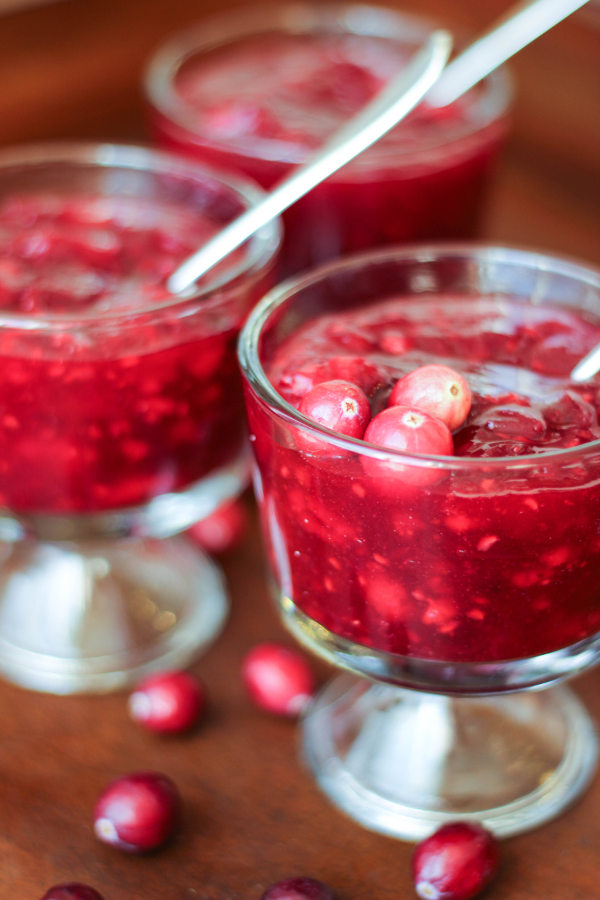 Three clear class bowls are full of crimson cranberry raspberry sauce. They have small spoons in them and a garnish of fresh cranberries.