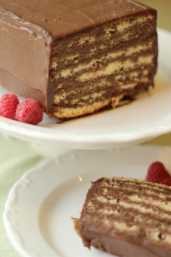 A beautiful, rich chocolate dobos torte sits on a white plate next to a few raspberries. The first slice is cut off and sitting on a plate next to the cake.