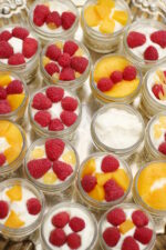 A tray is full of easy no bake fruit tarts covered in raspberries and peaches. The tarts are in tiny mason jars.