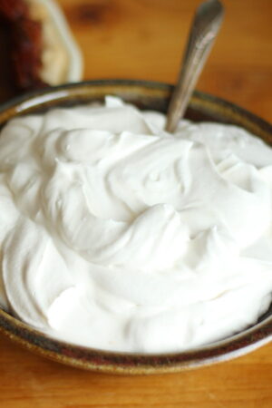 Brown pottery bowl of homemade whipped cream. The bowl is on a wooden table with a silver spoon in it.
