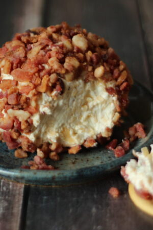 Close up image of a big smoked gouda cheese ball. The ball has been rolled in cooked bacon and you can see one slice off the end. It is sitting on a blue pottery plate.
