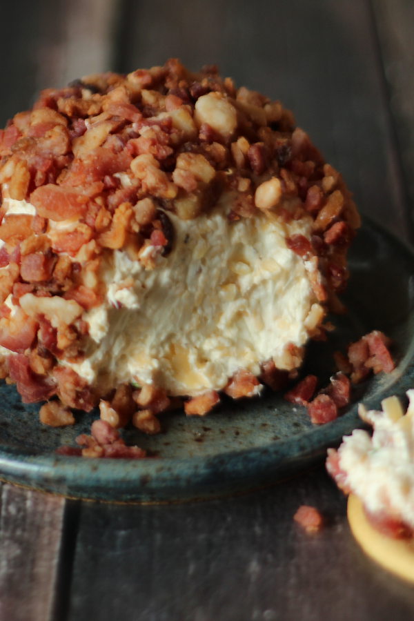 Close up image of a big smoked gouda cheese ball. The ball has been rolled in cooked bacon and you can see one slice off the end. It is sitting on a blue pottery plate.