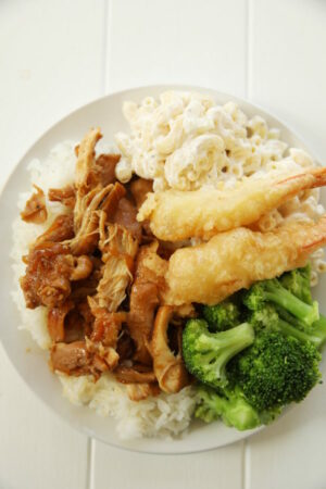 A big white plate is loaded up with rice covered in huli huli chicken. There is a big scoop of Mac Salad and a heap of broccoli with two tempura shrimp on top.