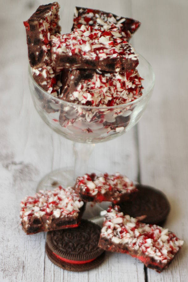 A stemmed glass is full of pieces of peppermint Oreo bark. There are additional pieces on the table with Oreo cookies.