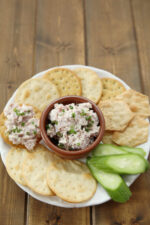 A platter of crackers and cucumbers has a dish of deviled ham spread in the middle waiting to be enjoyed.