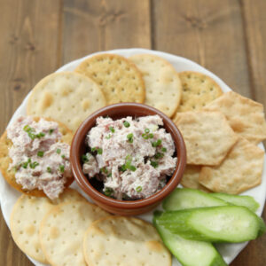 A platter of crackers and cucumbers has a dish of deviled ham spread in the middle waiting to be enjoyed.