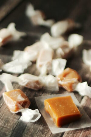 A dark wood table is covered with wrapped caramels. One caramel square is unwrapped ready to taste!