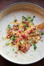 A big pot of loaded baked potato soup sits ready to eat with a wooden serving spoon in it. You can see piles of crumbled bacon, green onions, and cheddar cheese on top.