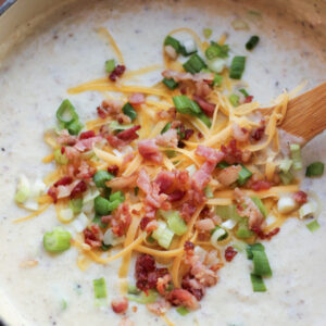 A big pot of loaded baked potato soup sits ready to eat with a wooden serving spoon in it. You can see piles of crumbled bacon, green onions, and cheddar cheese on top.