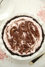 A delicious candy cane ice cream pie is in a white plate. You can see swirls of chocolate sauce on top and a crunchy oreo crust at the edge.