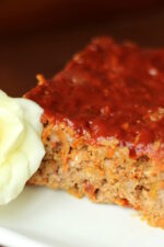 A tender loaf of meatloaf is sliced on a white plate. You can see the edge of a white rose next to the meatloaf.
