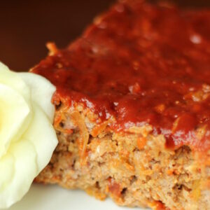 A tender loaf of meatloaf is sliced on a white plate. You can see the edge of a white rose next to the meatloaf.
