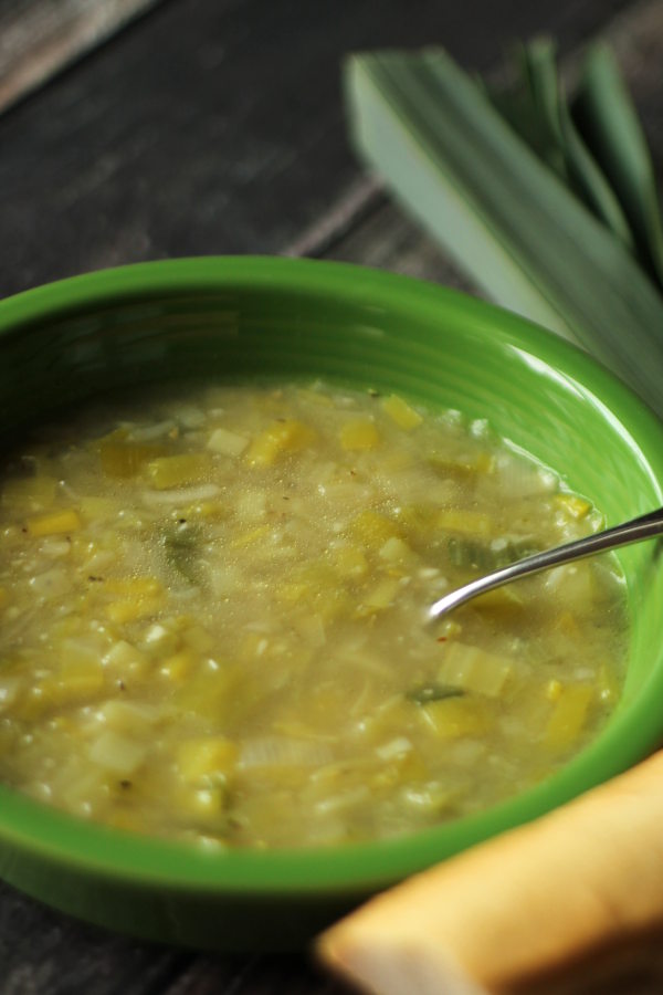 A green bowl of leek soup sits on a table. The soup is full of chicken broth and sautéed leeks.