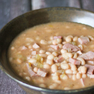 Brown pottery bowl full of ham and bean soup. You see a rich broth, white navy beans and big chunks of ham in the bowl.