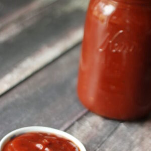 A mason jar full of spicy ketchup sits on a wooden table next to a dish of the ketchup ready for dipping.
