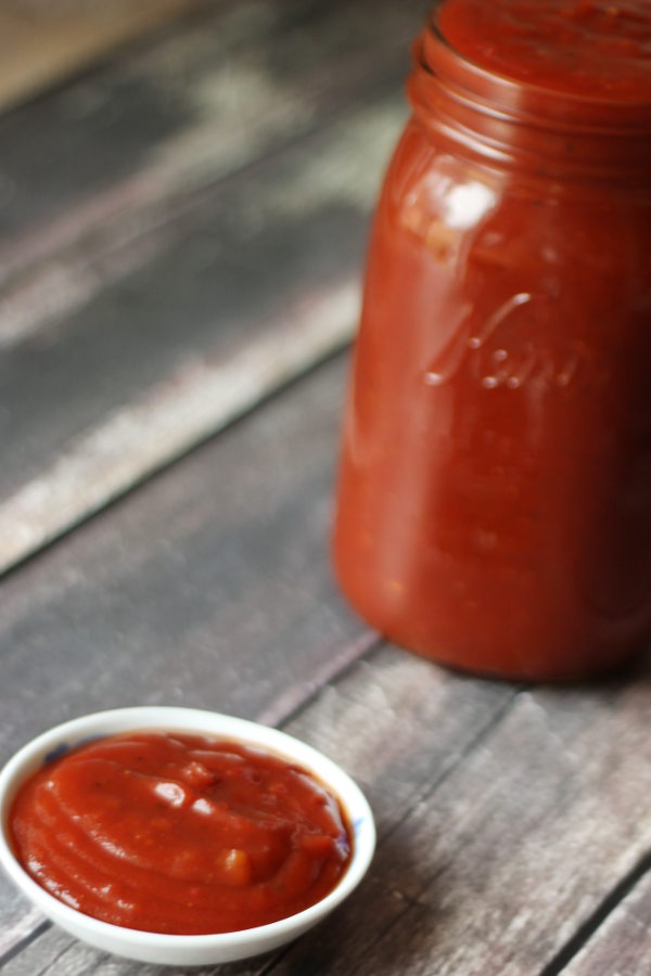 A mason jar full of spicy ketchup sits on a wooden table next to a dish of the ketchup ready for dipping.