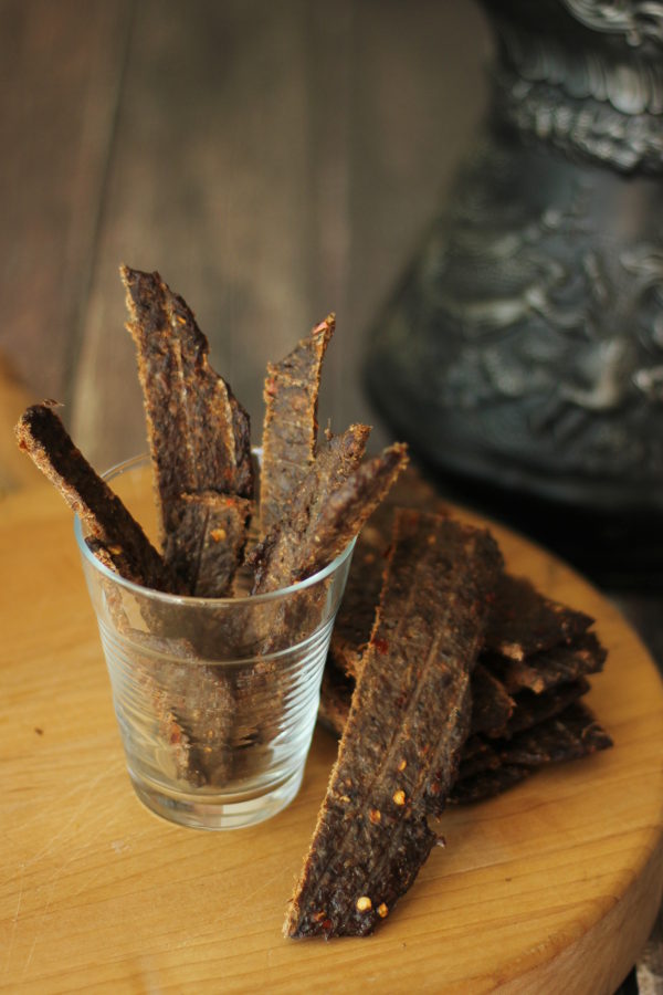 Beef jerky made with ground meat sits in a clear glass cup on a cutting board. More strips of jerky are laid next to it. Behind the jerky is a black pitcher.
