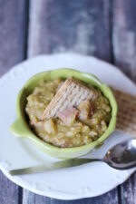 A green bowl of thick split pea soup sits on a white plate. There are big chunks of ham in the soup and a cracker on top.