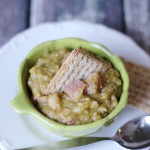 A green bowl of thick split pea soup sits on a white plate. There are big chunks of ham in the soup and a cracker on top.