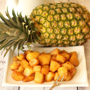 Chunks of pineapple roasted with brown sugar, cinnamon and butter. The pineapple is piled on a white plate in front of a whole pineapple.