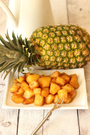 Chunks of pineapple roasted with brown sugar, cinnamon and butter. The pineapple is piled on a white plate in front of a whole pineapple.