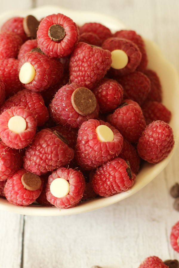 A white bowl is full of fresh raspberries. Each berry has a white chocolate chip, or a semi-sweet chocolate chip or a milk chocolate chip tucked inside of it.