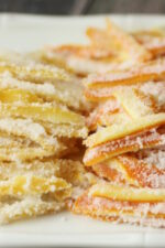 A white platter is stacked high with bright yellow strips of candied lemon peel and bright orange strips of candied orange peel. Both stacks of candied citrus peel glisten with sugar.