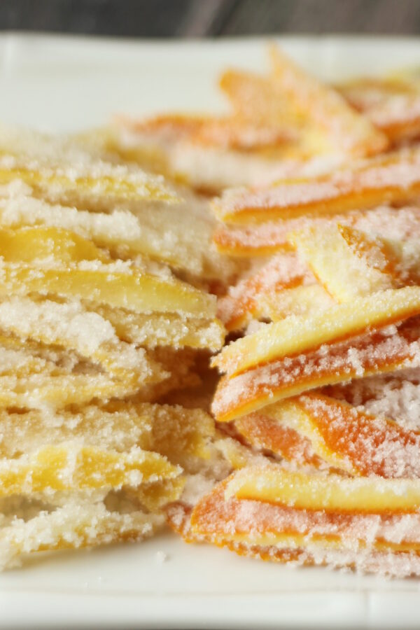 A white platter is stacked high with bright yellow strips of candied lemon peel and bright orange strips of candied orange peel. Both stacks of candied citrus peel glisten with sugar.