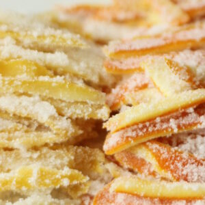 A white platter is stacked high with bright yellow strips of candied lemon peel and bright orange strips of candied orange peel. Both stacks of candied citrus peel glisten with sugar.