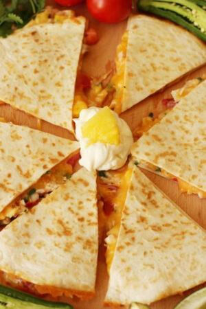 A big pineapple chicken quesadilla sits cut into six pieces on the cutting board. There is sour cream in the middle ready for dipping and there is pineapple, tomato, and jalapeno around the quesadilla.