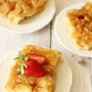Three slices of creme brulee french toast casserole on white plates. One slice has strawberries on top.