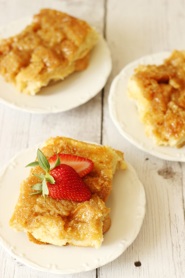 Three slices of creme brulee french toast casserole on white plates. One slice has strawberries on top.