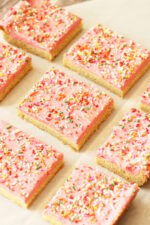Fresh baked sugar cookie bars are laid out in a grid on parchment paper. The bars are frosted with pink raspberry buttercream and covered with brite sprinkles.