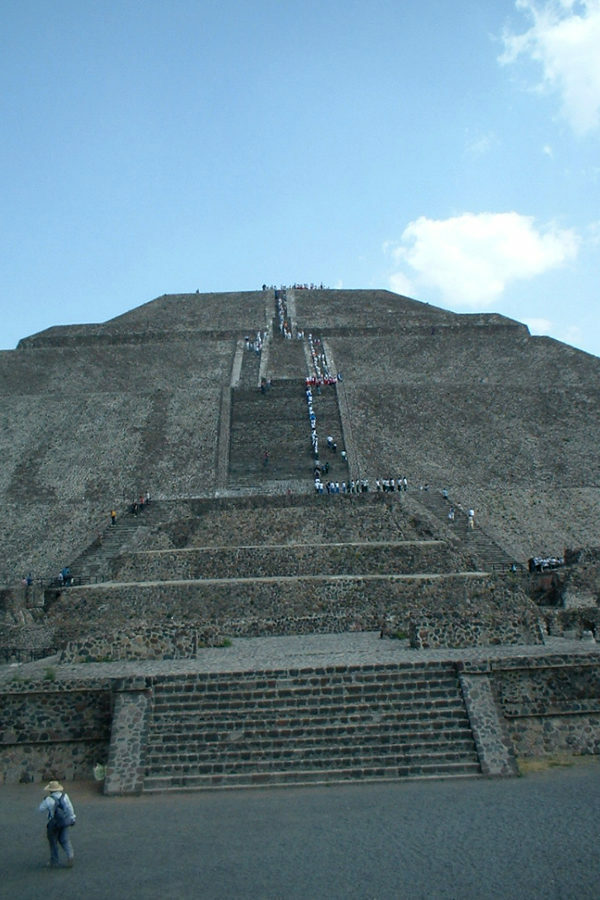 Teotihuacan near Mexico city stands tall against a blue sky with white puffy clouds. You can see a few people climbing it.