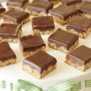 Oatmeal cookie bars are arranged on a white cake stand. Each bar is covered with a thick layer of rich chocolate frosting.