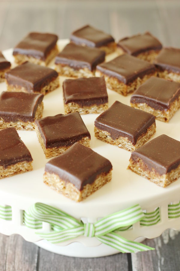 Oatmeal cookie bars are arranged on a white cake stand. Each bar is covered with a thick layer of rich chocolate frosting.