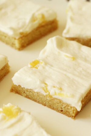 A tray is lined with parchment paper and squares of sugar cookie bars sit on the paper. You can see the coconut-pineapple buttercream frosting on top of the bars. There are chunks of pineapple visible too.