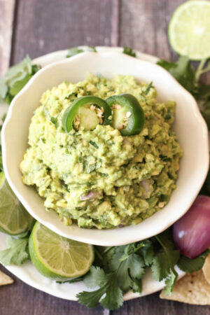 A white bowl is mounded high with chunky, fresh, guacamole. It is garnished with a few jalapenos and surrounded with limes, cilantro and some shallots.